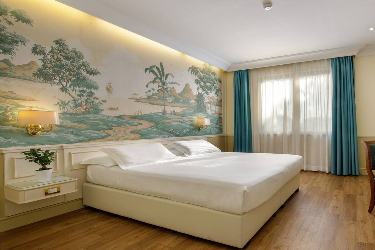 A hotel room with a large bed, a desk with a chair, a desk lamp, a wall-mounted TV, and a painting on the wall.