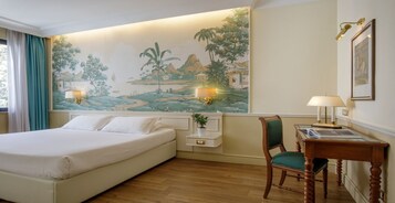 Una camera d'albergo con un letto grande, una scrivania con una sedia, un tavolino con una lampada e un murale raffigurante un paesaggio tropicale.