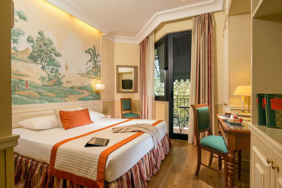 Habitación de hotel con cama, escritorio, silla, espejo y balcón con cortinas.