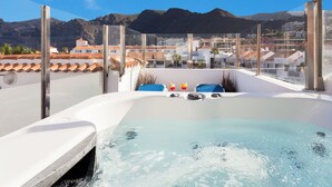 Una vasca idromassaggio con vista sulle montagne, circondata da un recinto in vetro, e una zona piscina con lettini e un tavolo.