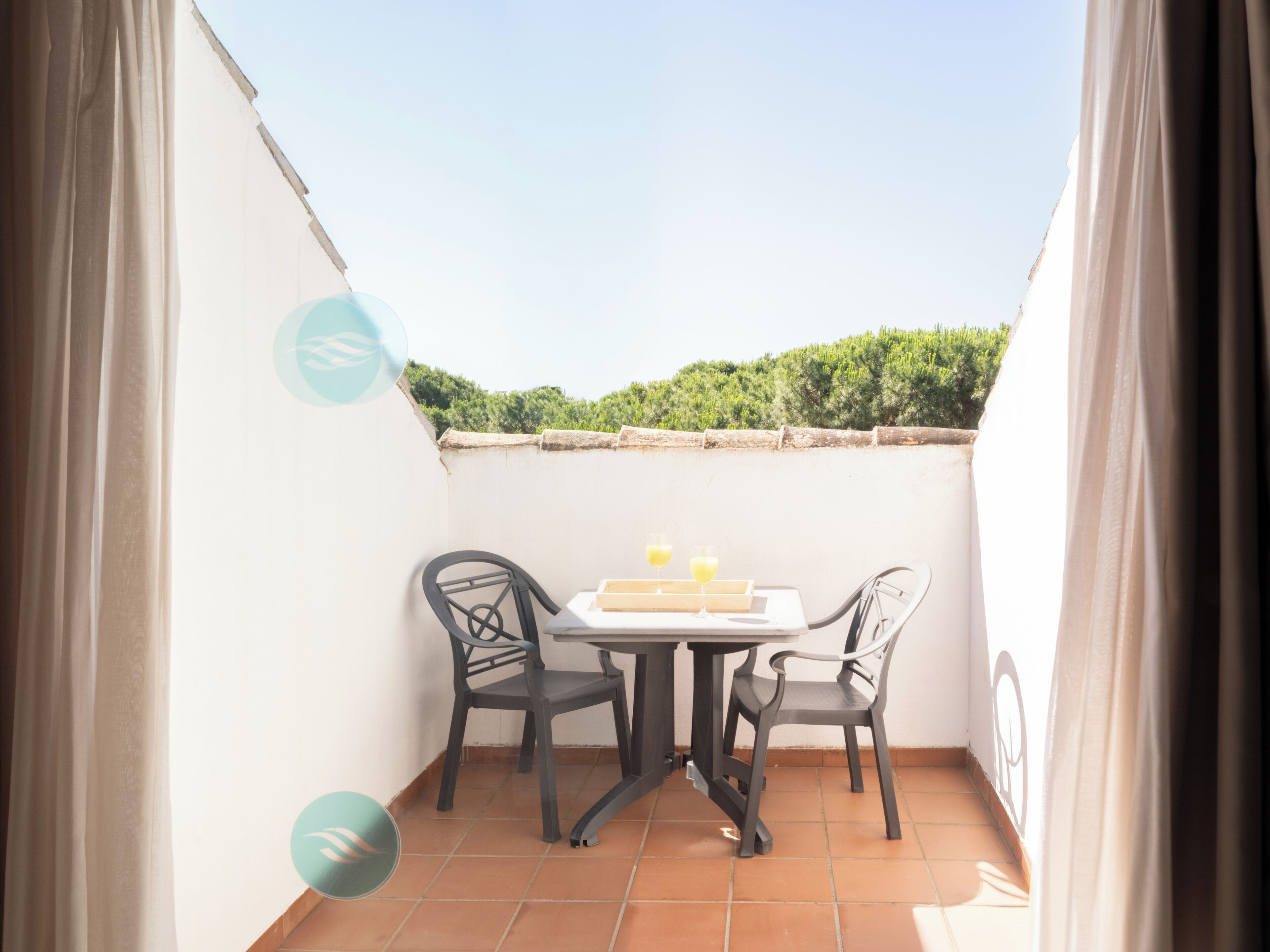A small outdoor balcony with a table and two chairs, a tiled floor, and a view of greenery in the background.
