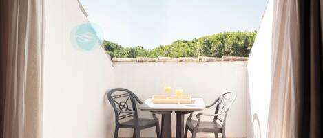 A small outdoor balcony with a table and two chairs, a tiled floor, and a view of greenery in the background.