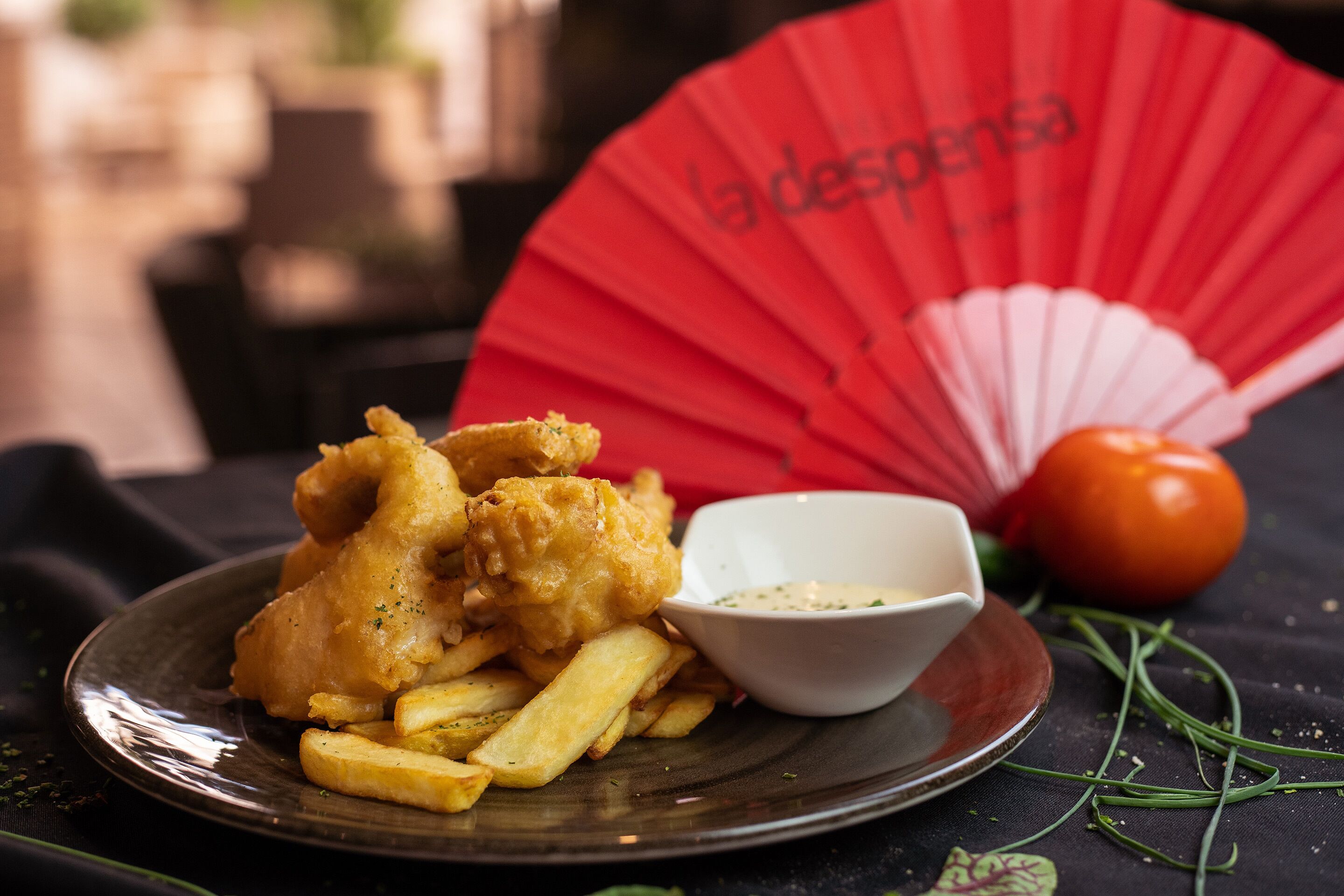 Un plato de pescado con patatas fritas, un recipiente pequeño de salsa, un tomate y un abanico rojo con texto.