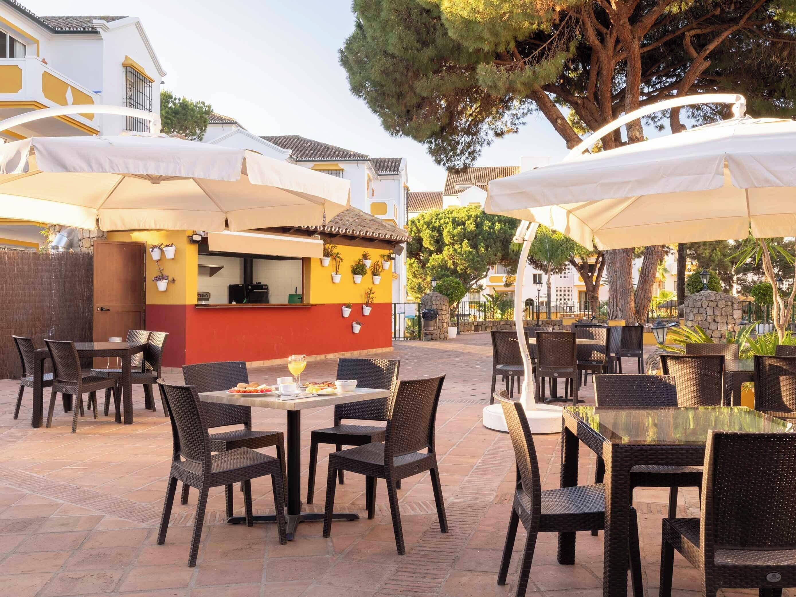 An outdoor patio with wicker tables and chairs, umbrellas, and a food service area.