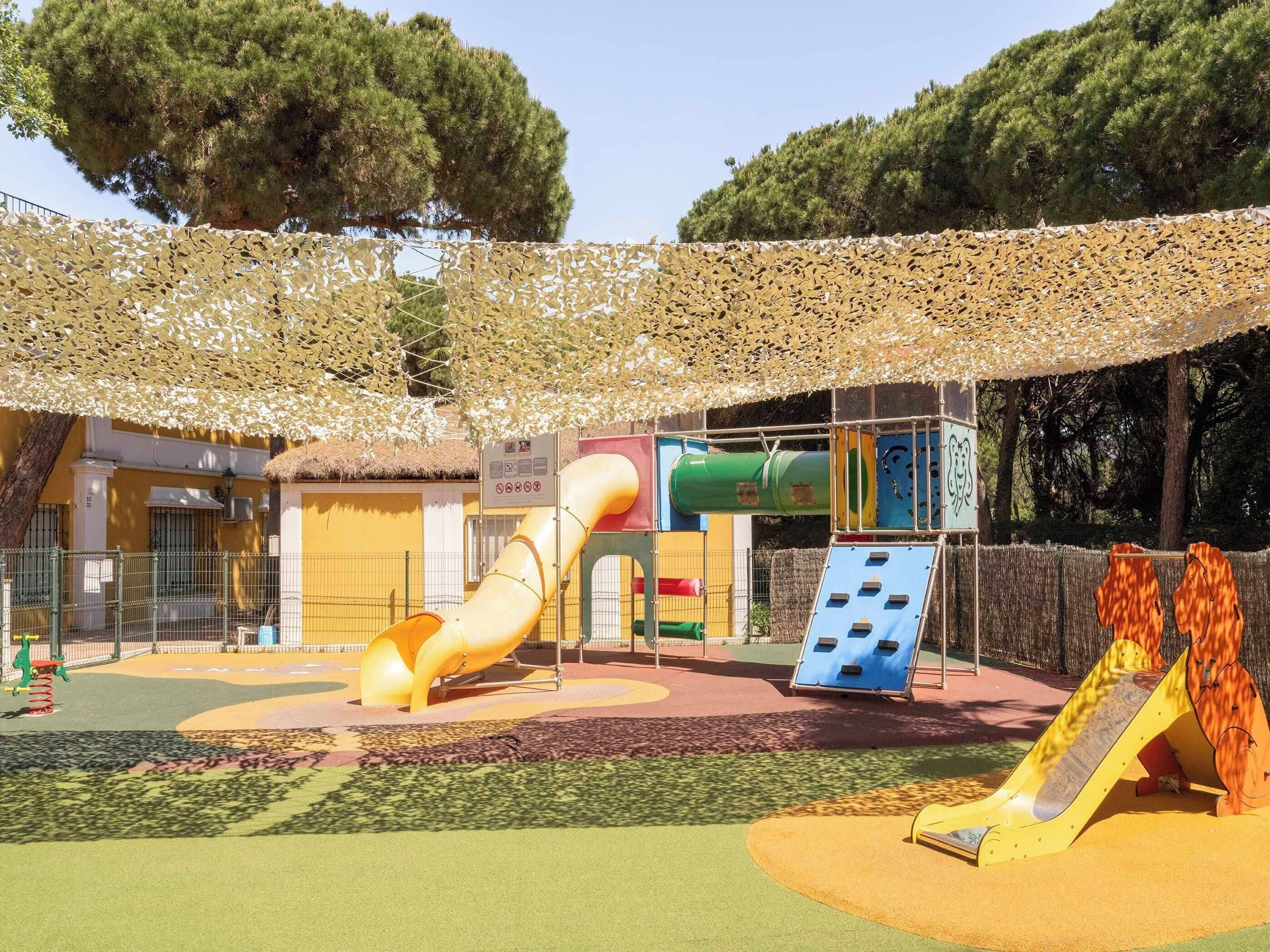 Un parque infantil con toboganes y estructuras para escalar, rodeado por una cerca y árboles.