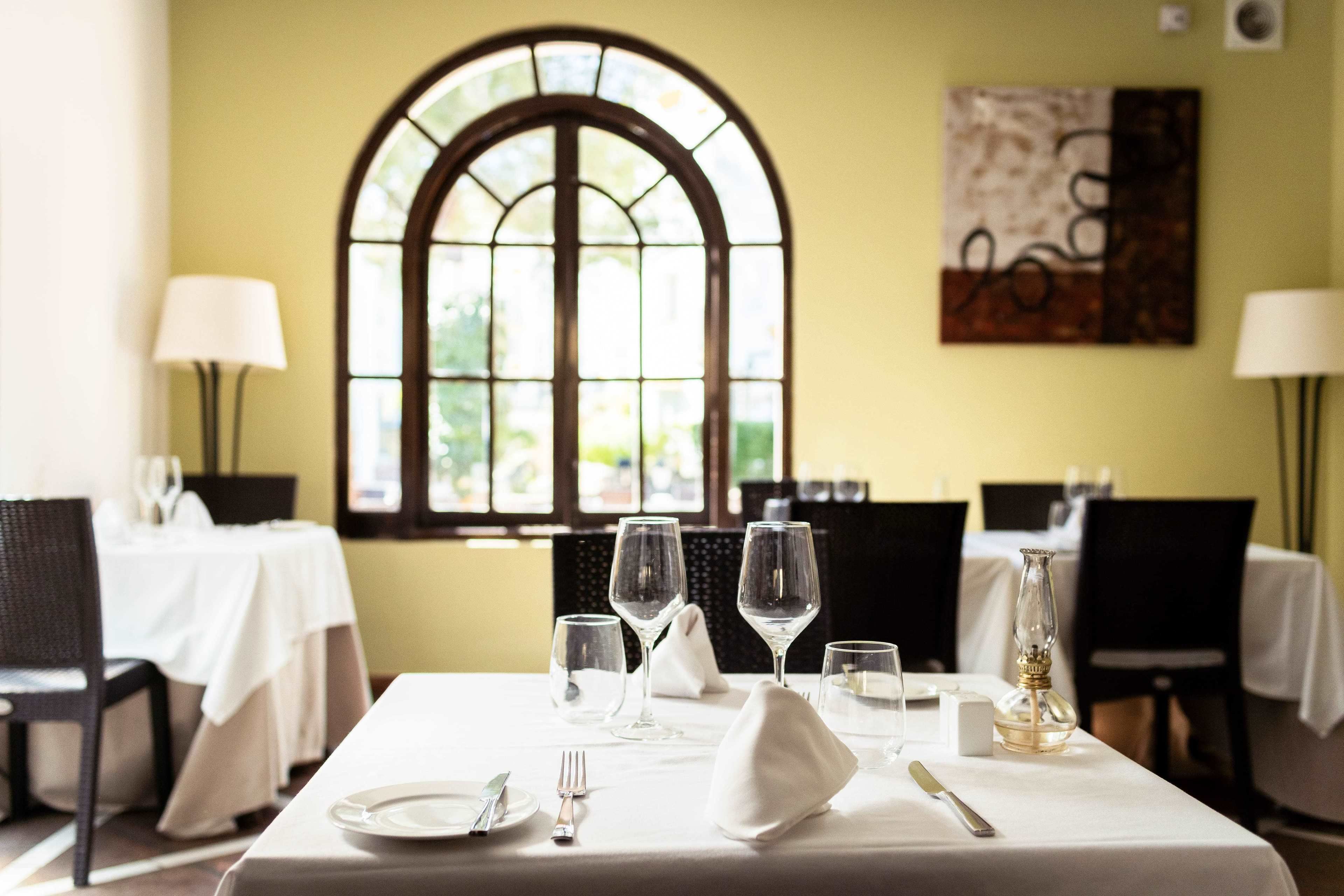 Tavola da pranzo apparecchiata con tovaglia bianca, piatti, posate e bicchieri da vino, con una grande finestra ad arco e un quadro appeso al muro.