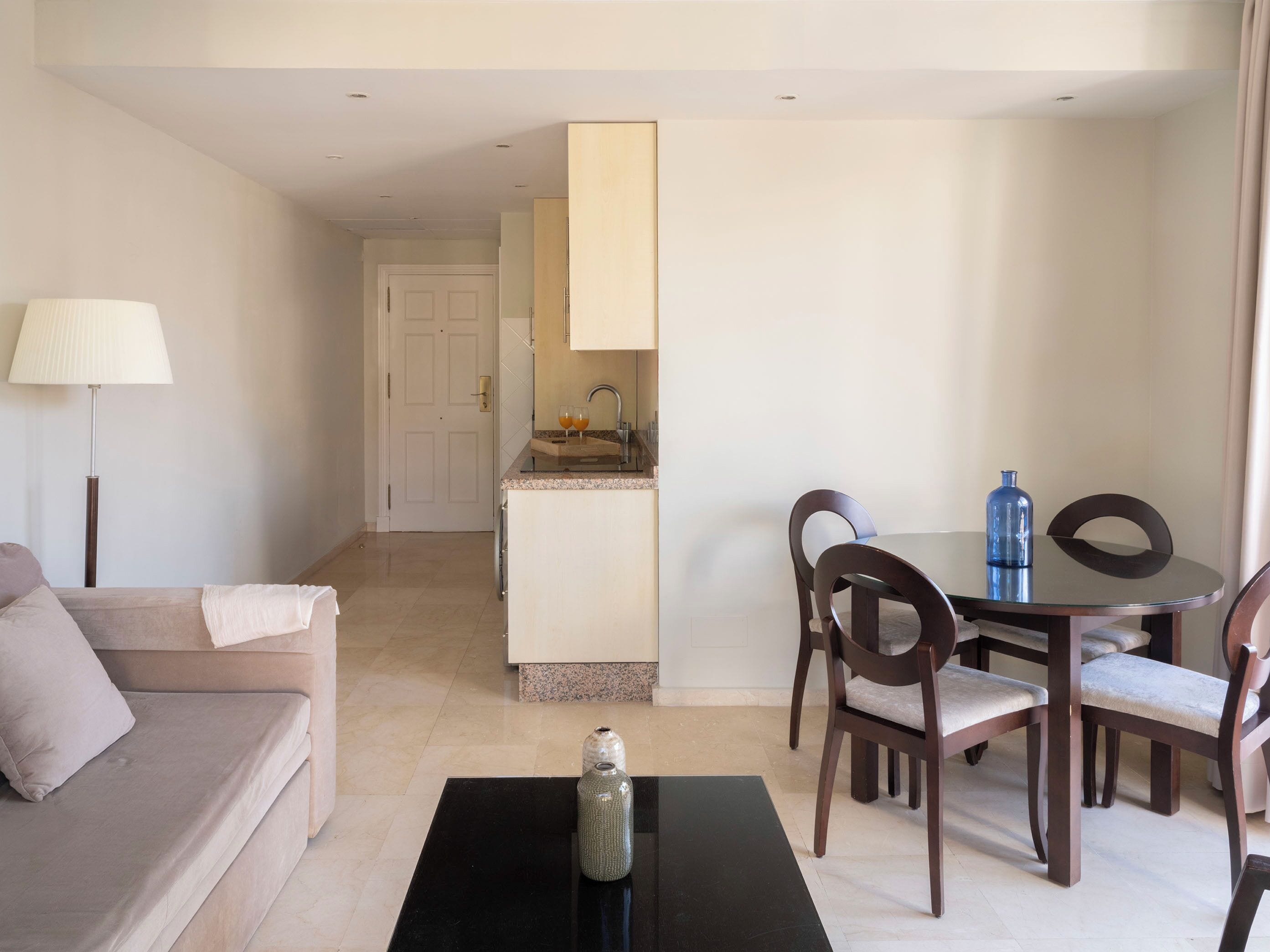 A hotel room with a sofa, a dining table with chairs, a kitchen area, and a lamp.