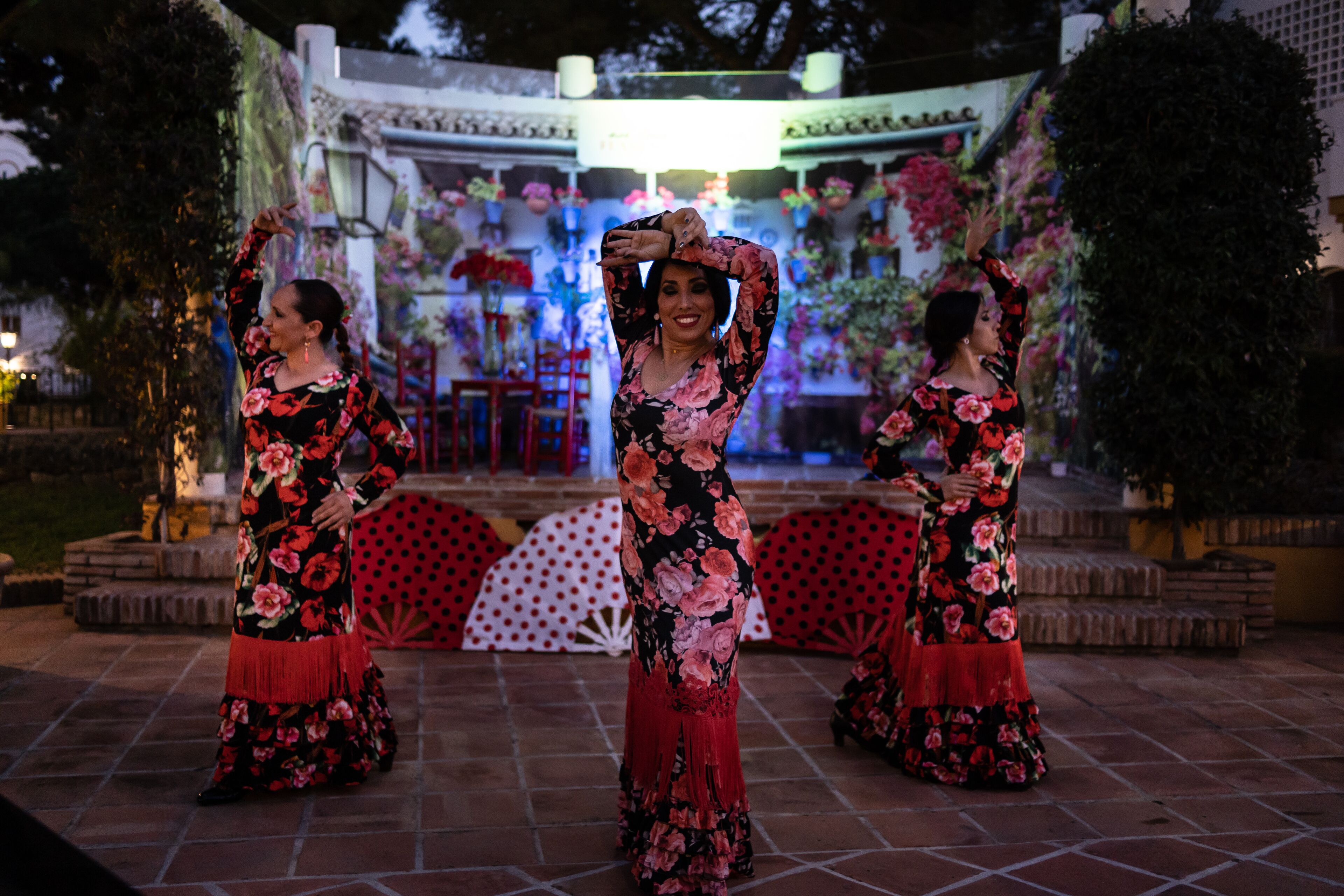 Un patio con un escenario decorado con flores y plantas, y tres mujeres con vestidos florales actuando.