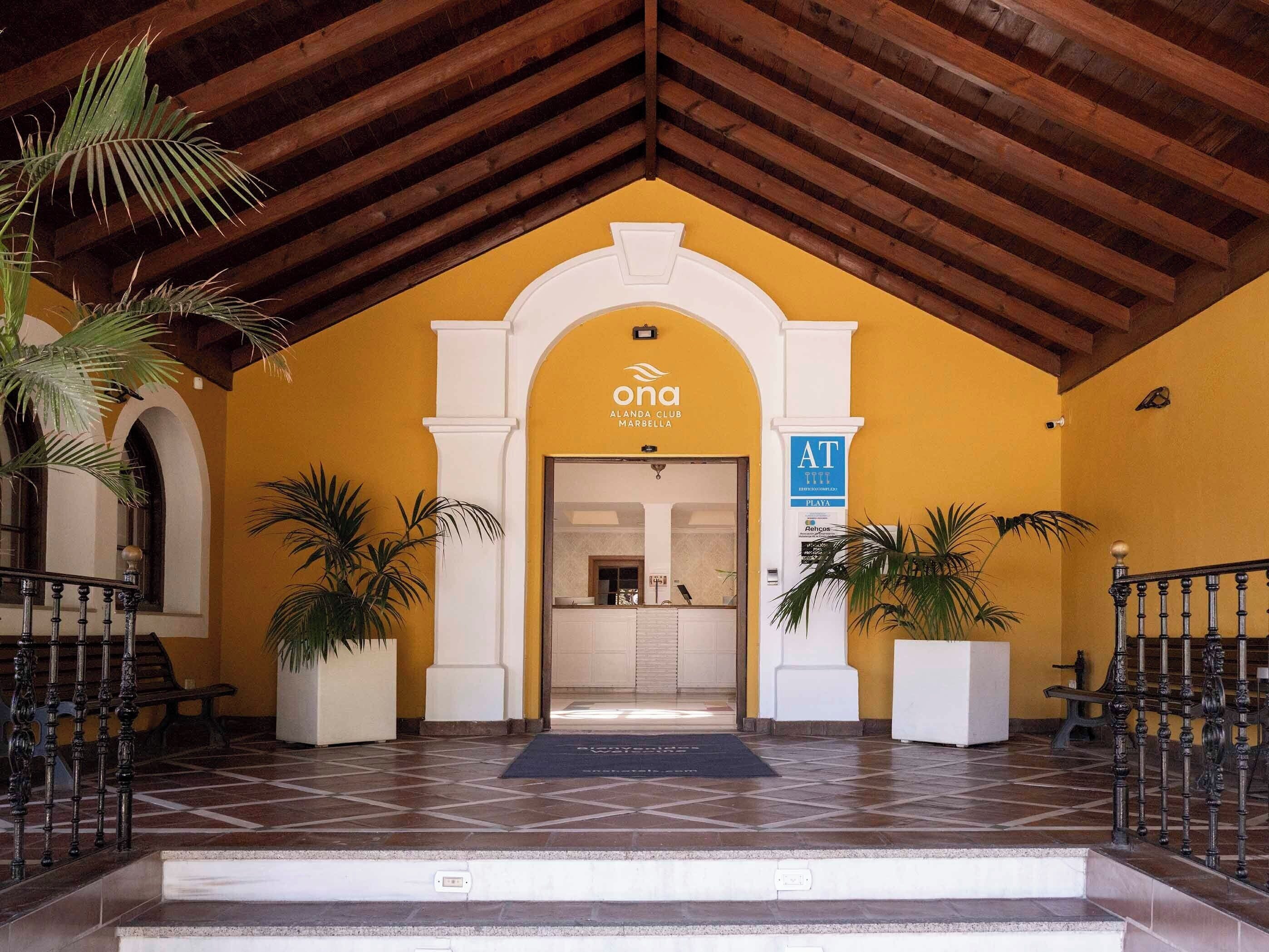 Ein Hotel Eingang mit gelber Fassade, Holzbalkendecke und einem Schild mit der Aufschrift 'ona ALANDA CLUB MARBELLA'.