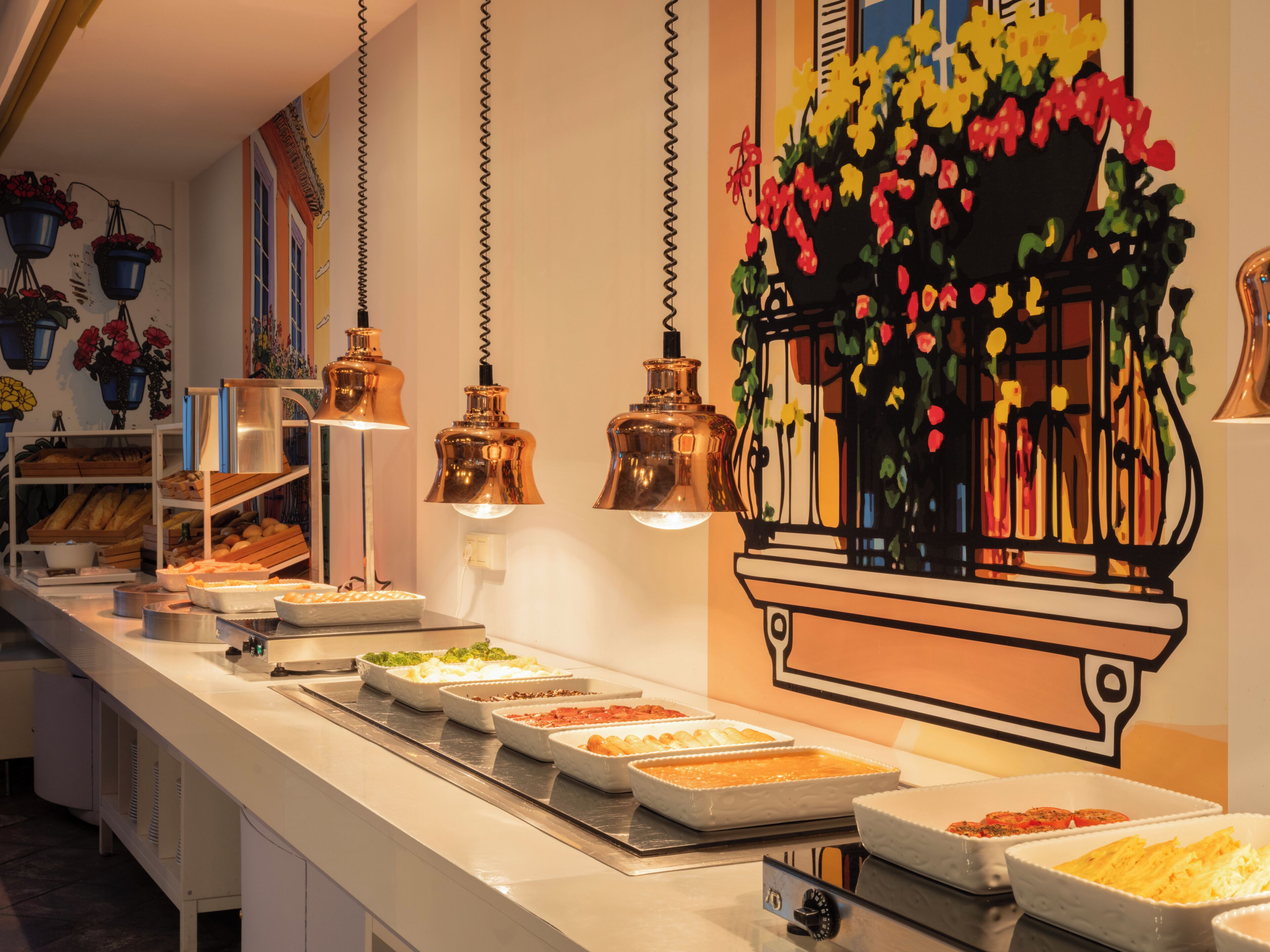A buffet setup with various food items, hanging pendant lights, and a decorative wall with a floral motif.