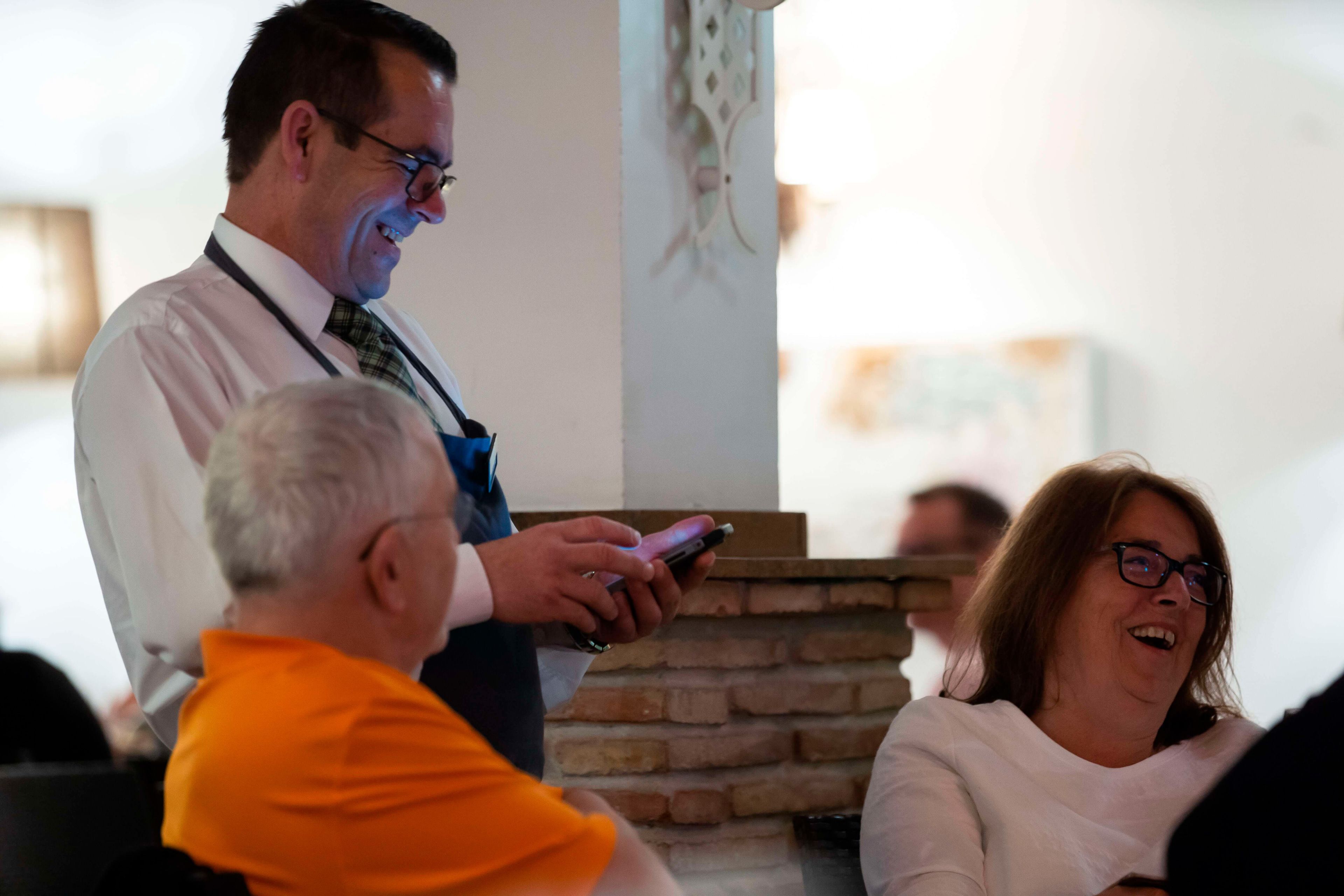 Een man in een wit overhemd en das vasthoudend een smartphone, staand in een kamer met een bakstenen muur en een decoratieve lamp.