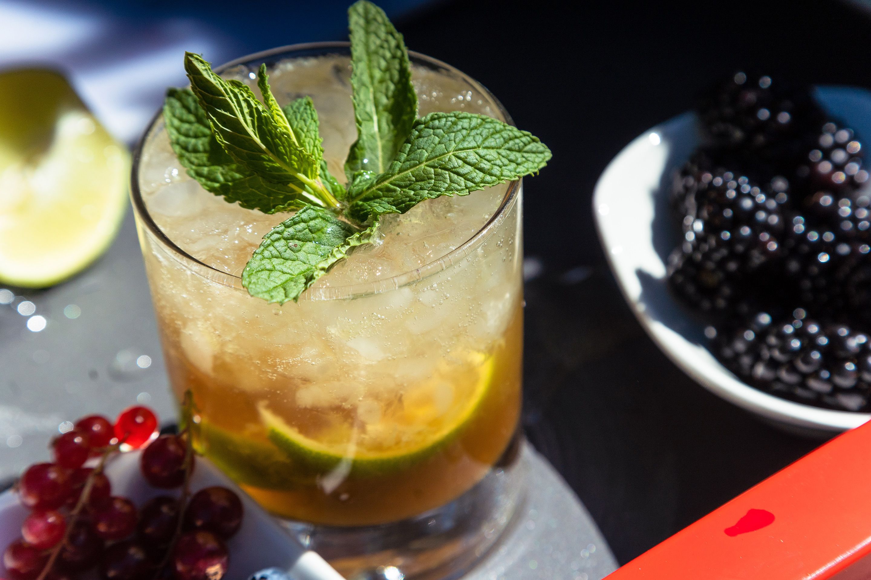Un verre de boisson glacée garni de feuilles de menthe et d’une tranche de citron vert, avec un bol de mûres en arrière-plan.