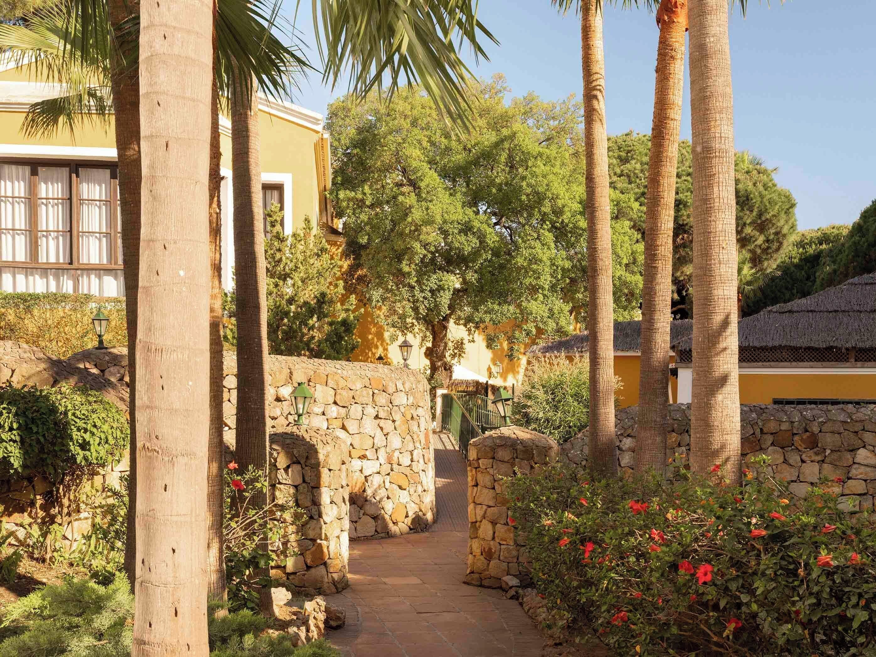 A stone pathway leading to a building with a yellow facade, surrounded by palm trees and flowering plants.