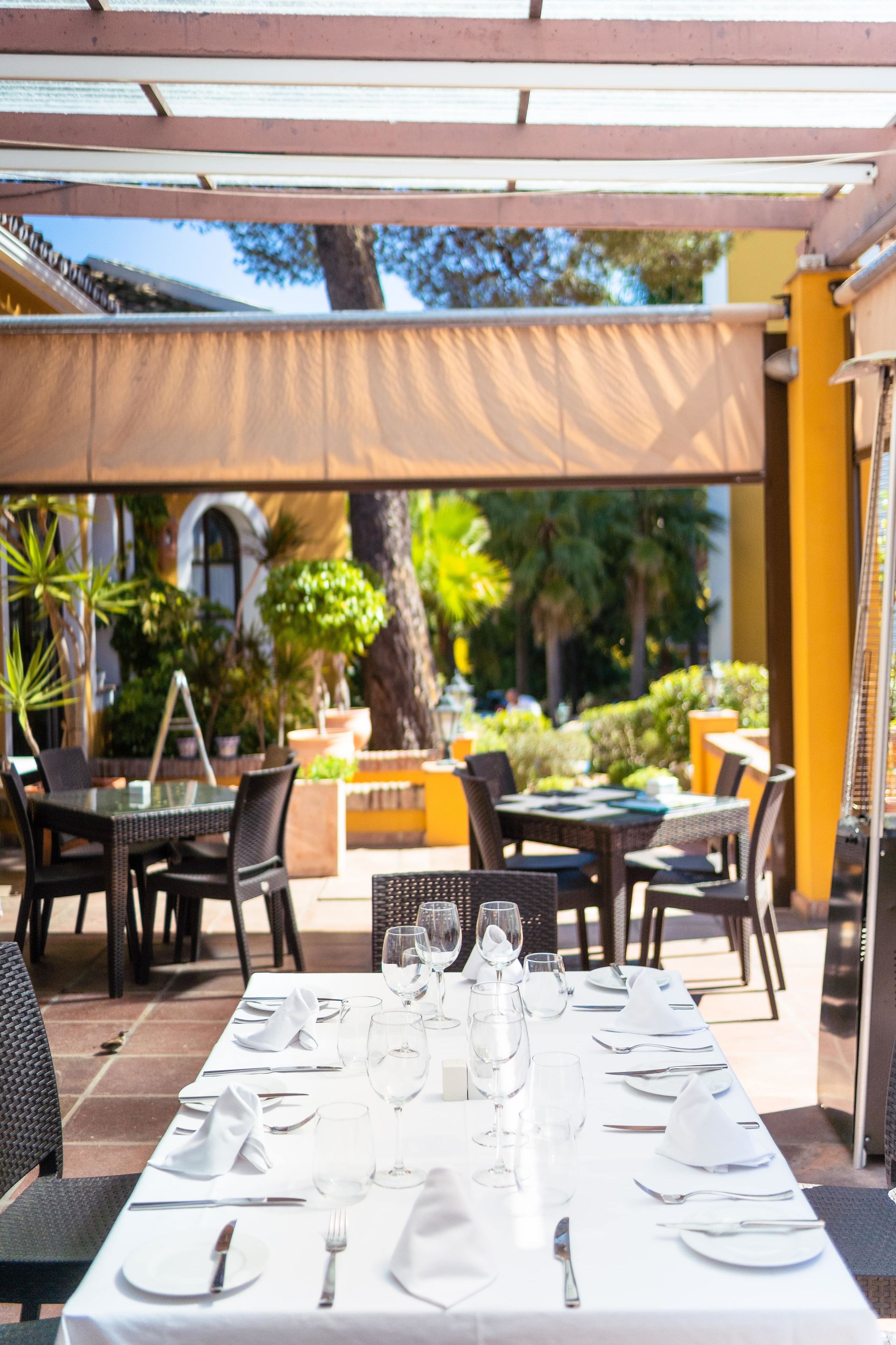 An outdoor dining area with tables set for two, surrounded by wicker chairs and a pergola providing shade.