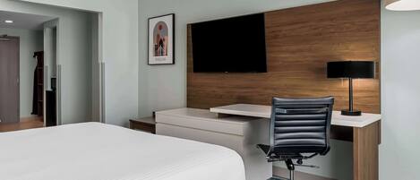 Habitación de hotel con una cama grande, un escritorio con silla, un televisor de pantalla plana en la pared y un cuadro con una vista urbana.