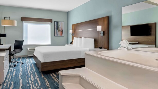 A hotel room with a bed, a desk, a chair, a large window, a painting, a headboard, a TV, and a bathtub. - Best Western Executive Inn (Seagoville)