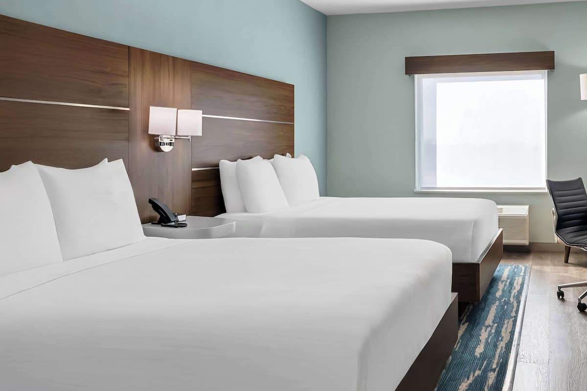 A hotel room with two beds, a wooden headboard, a bedside lamp, a window with blinds, and a chair.