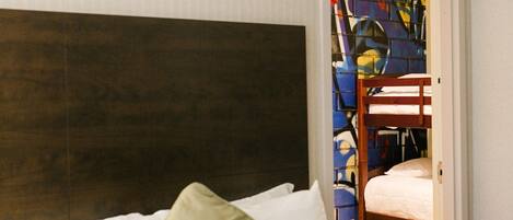 Une chambre d’hôtel avec un lit, une tête de lit en bois et un lit superposé visible à travers une porte ouverte.