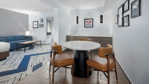 A modern living room with a blue sofa, a coffee table, and a round dining table with four chairs. There is a kitchen area with a sink and framed pictures on the wall.