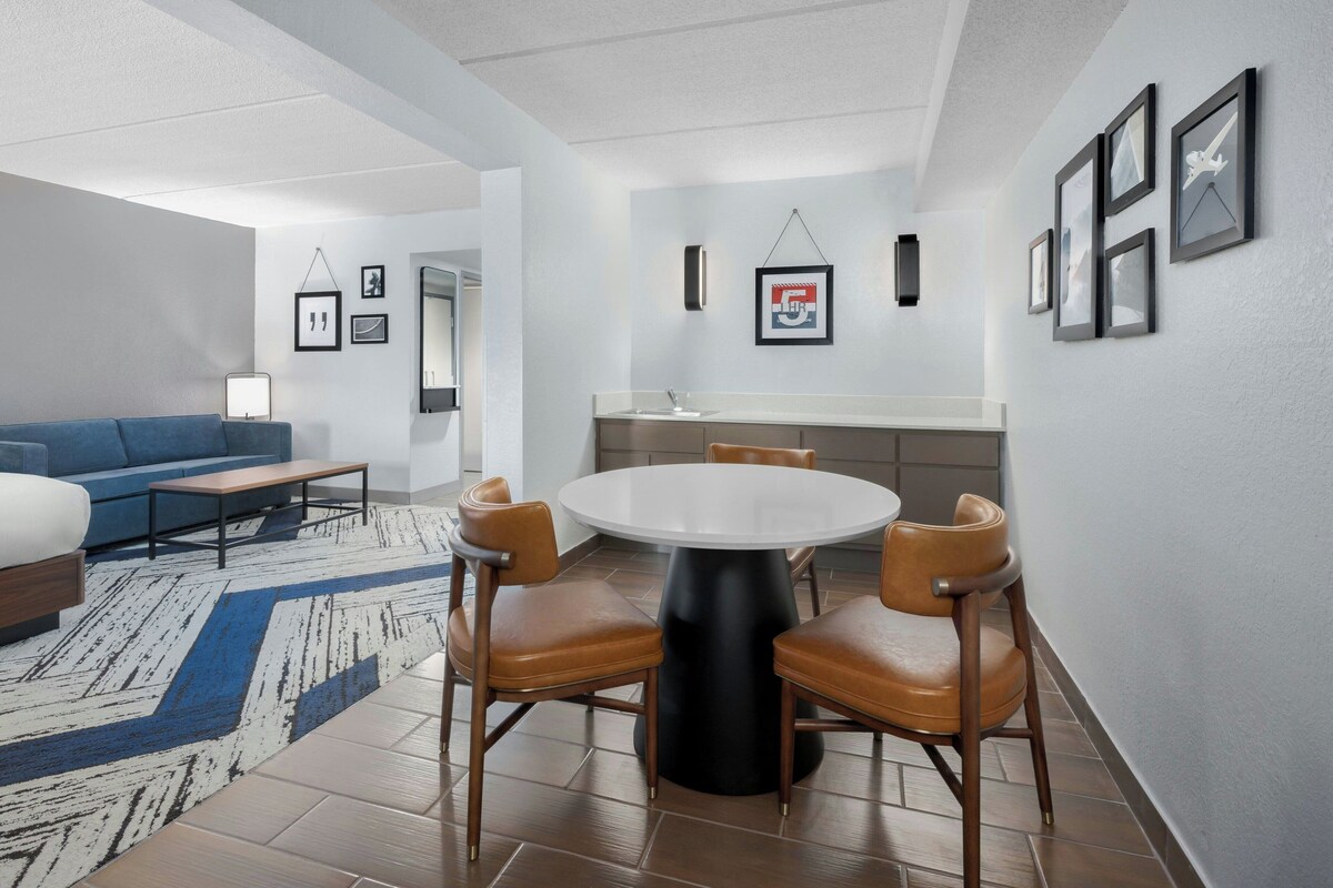 Een moderne woonkamer met een blauwe bank, een salontafel en een ronde eettafel met vier stoelen. Er is een keukenhoek met een spoelbak en afbeeldingen in lijsten aan de muur.