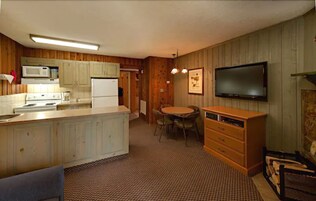 A compact kitchen with wooden cabinets, a microwave, a refrigerator, a dining table with chairs, and a television mounted on the wall.