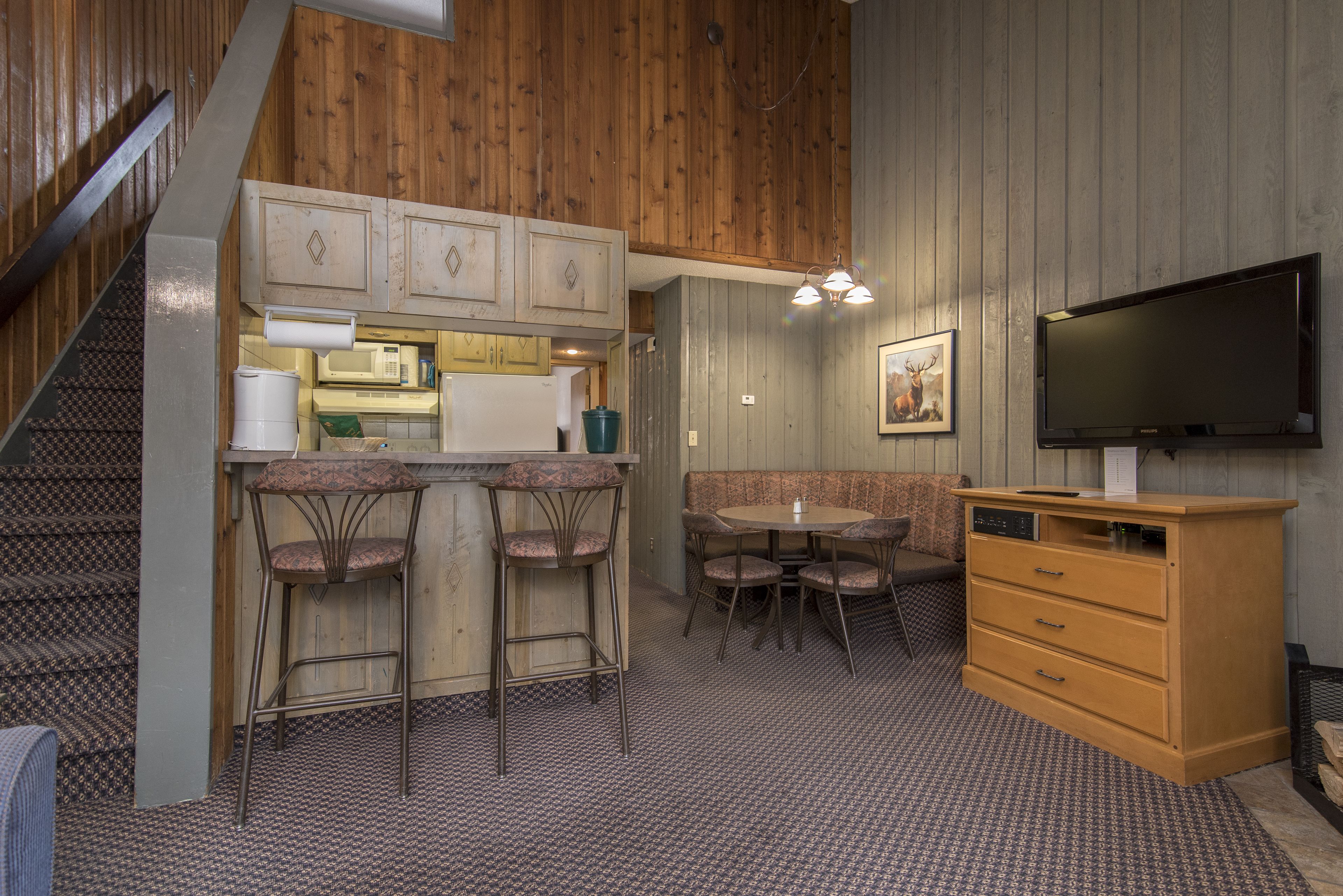 A compact kitchen with a microwave, a sink, and a bar area with stools. There is a television mounted on the wall and a wooden cabinet with drawers.