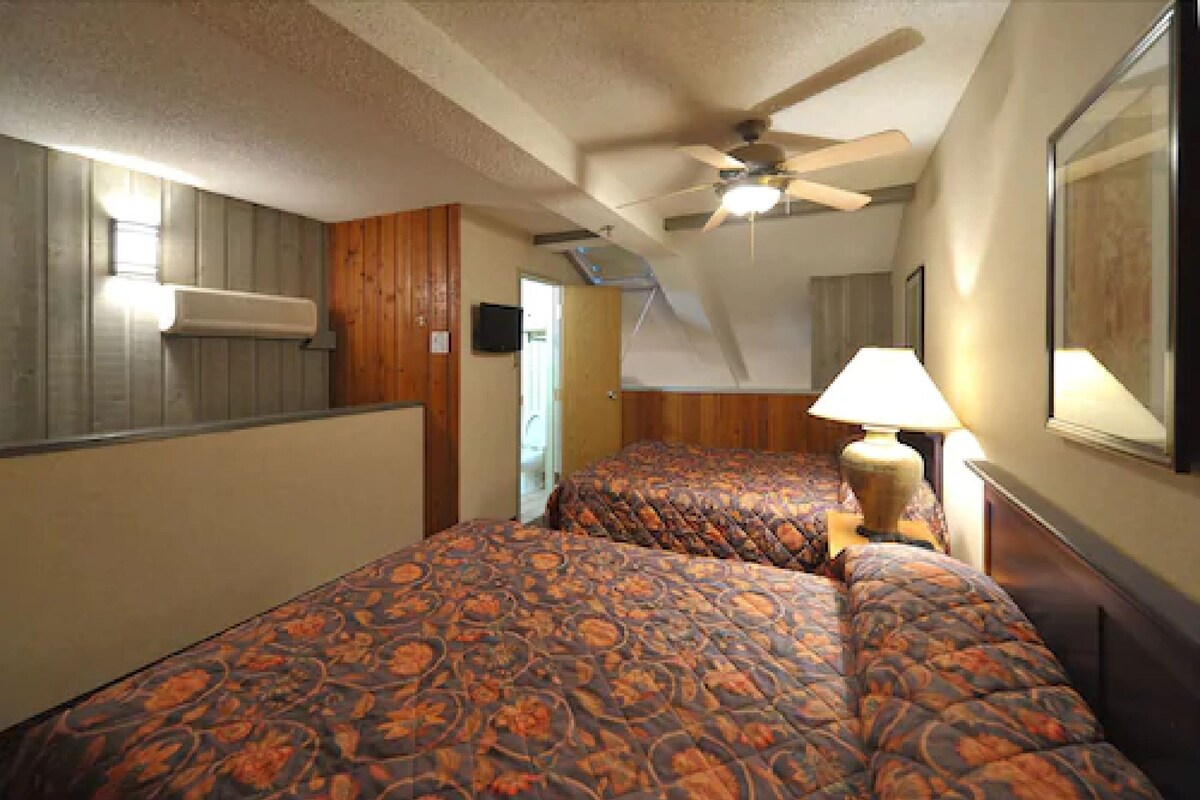 Habitación de hotel con dos camas, ventilador de techo, lámpara y un cuadro en la pared.