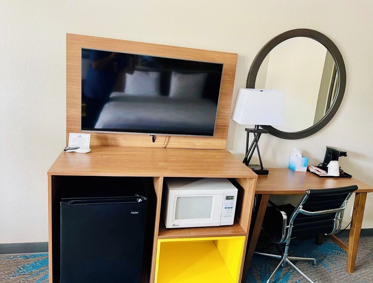 A hotel room with a wooden desk, a television, a microwave, a refrigerator, a chair, a lamp, and a mirror.