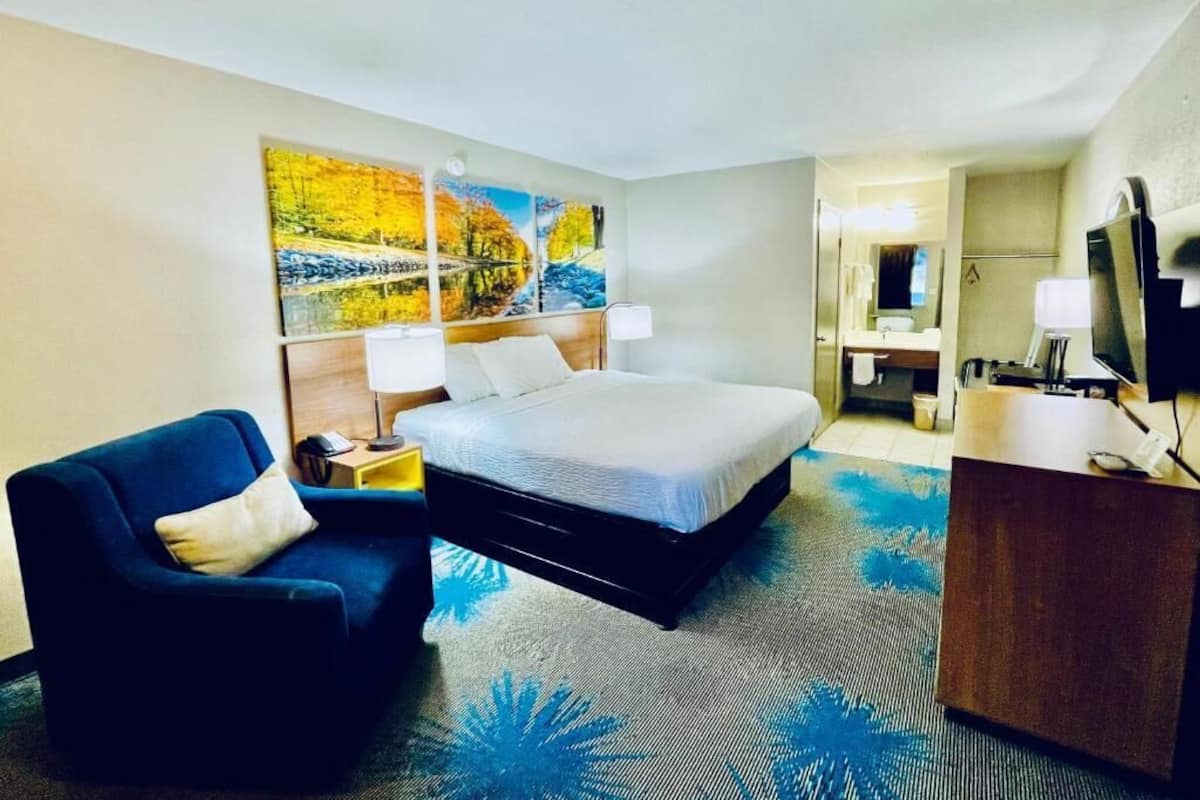 A hotel room with a bed, a chair, a desk, a mirror, and artwork on the wall.