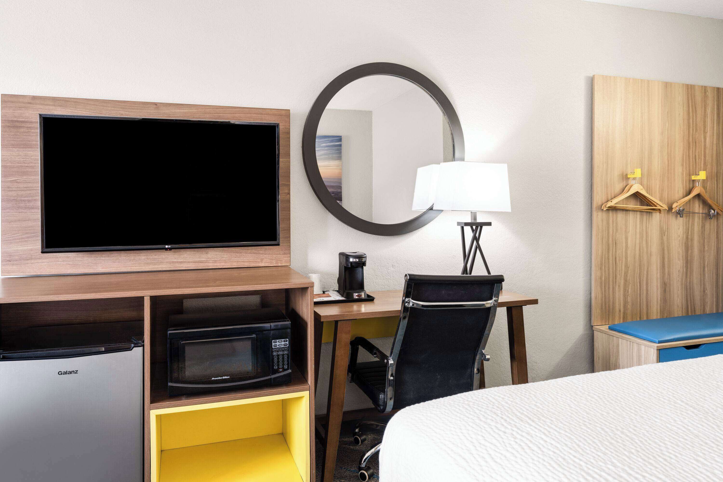 A hotel room with a flat-screen TV, a microwave, a desk with a lamp, a chair, and a bed.