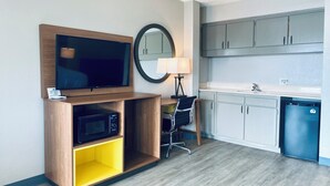 A modern hotel room with a flat-screen TV, a microwave, a chair, a desk, a lamp, a mirror, and a kitchenette with cabinets and a sink.