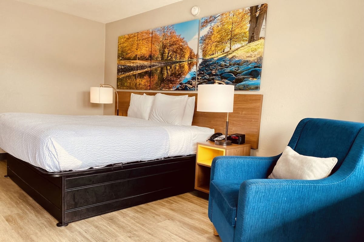 A hotel room with a bed, a blue armchair, a nightstand with a lamp, and two paintings on the wall.