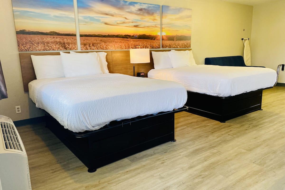 A hotel room with two beds, a large window with a sunset view, a heater, and a wall-mounted lamp.