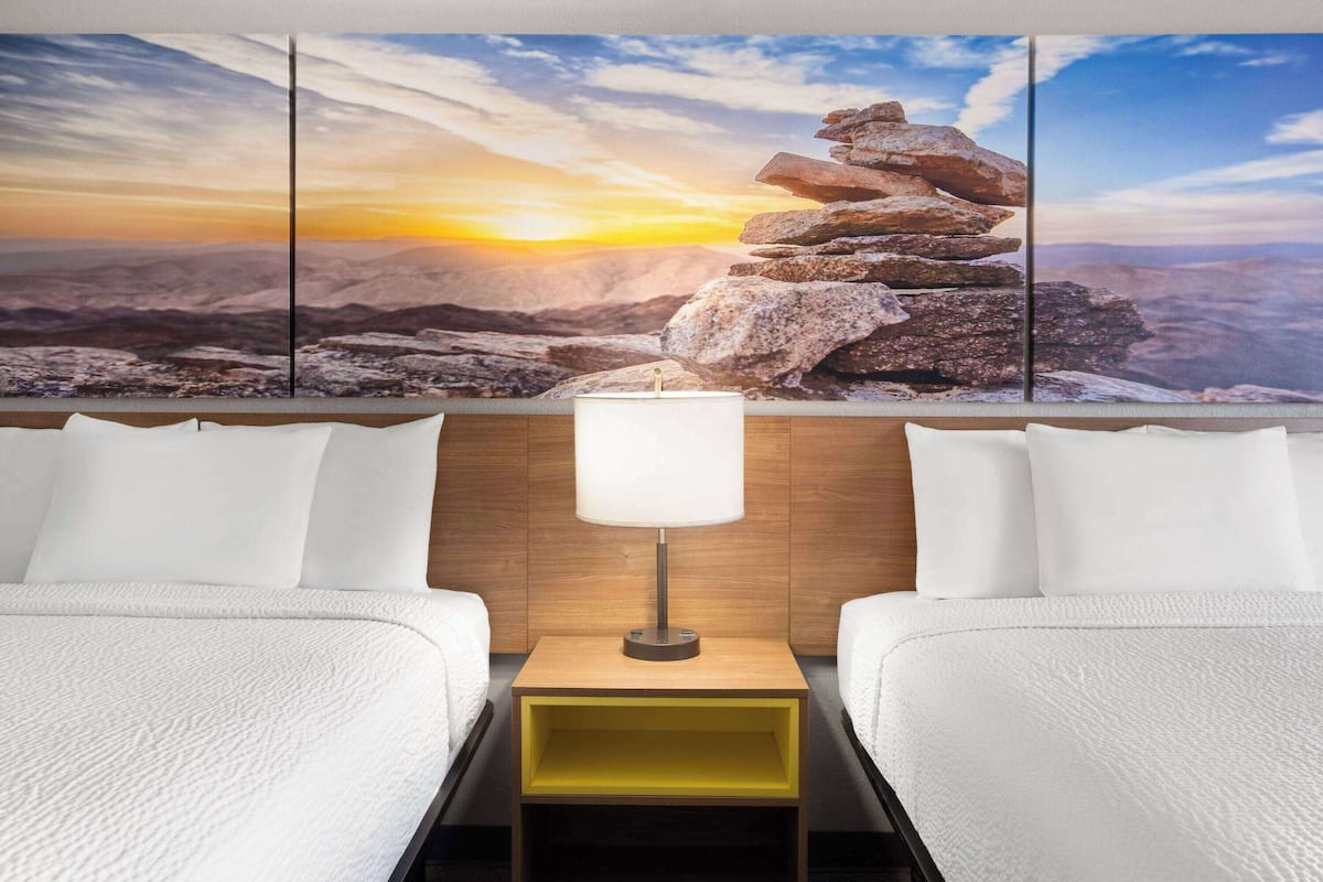 A hotel room with two beds, a wooden headboard, a nightstand with a lamp, and a large landscape painting above the beds.