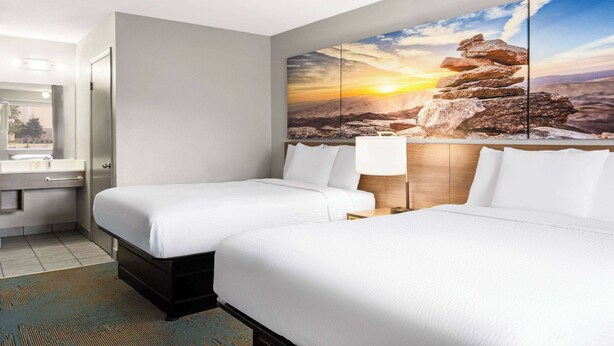 A hotel room with two beds, a large painting of a sunset over mountains, a bathroom with a sink and mirror, and a wooden headboard.