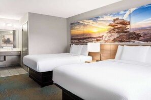 Ein Hotelzimmer mit zwei Betten, einem großen Gemälde mit Sonnenuntergang über Bergen, einem Badezimmer mit Waschbecken und Spiegel sowie einem Holzkopfteil.