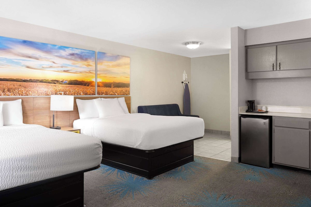 A hotel room with two beds, a kitchenette, and a large painting of a sunset over a field.