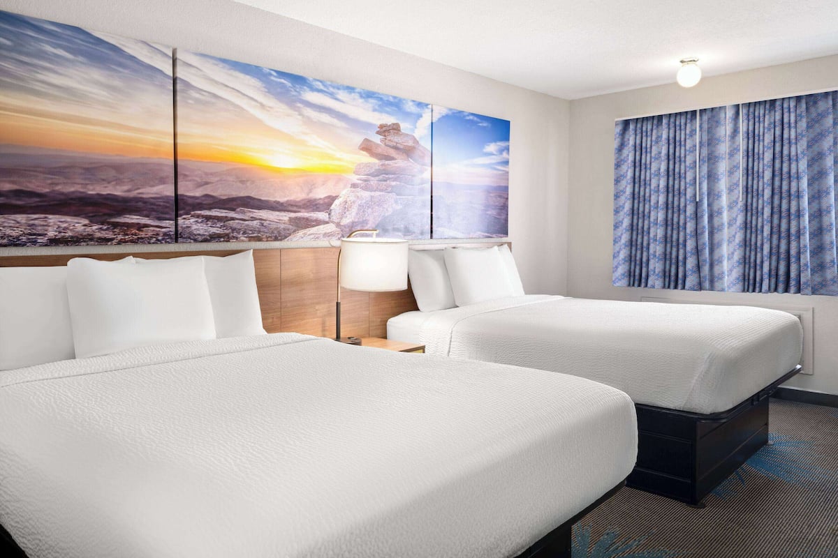 A hotel room with two beds, a large painting of a mountain landscape, a nightstand with a lamp, and a window with curtains.