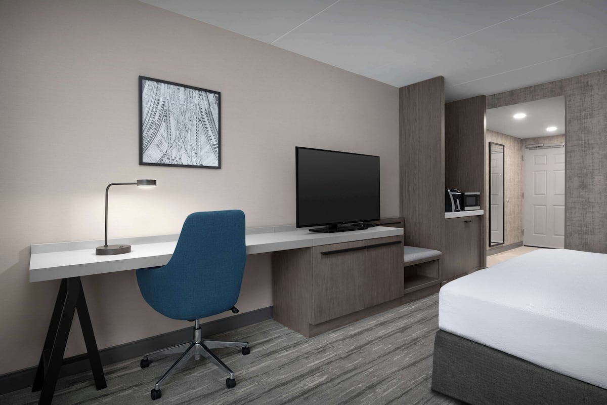 Ein Hotelzimmer mit einem Bett, einem Schreibtisch mit Stuhl, einem Fernseher und einem gerahmten Bild an der Wand.