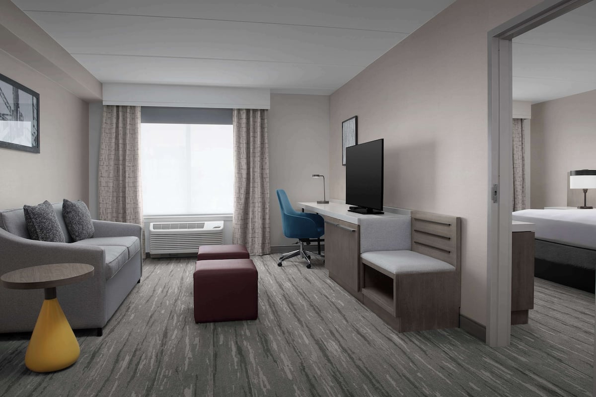 Ein modernes Hotelzimmer mit grauem Sofa, einem kleinen runden Tisch, einem blauen Bürostuhl, einem Fernseher, einem Schreibtisch und einem Bett.