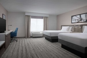 Habitación de hotel con dos camas, un escritorio con silla, un televisor y un amplio ventanal con cortinas.