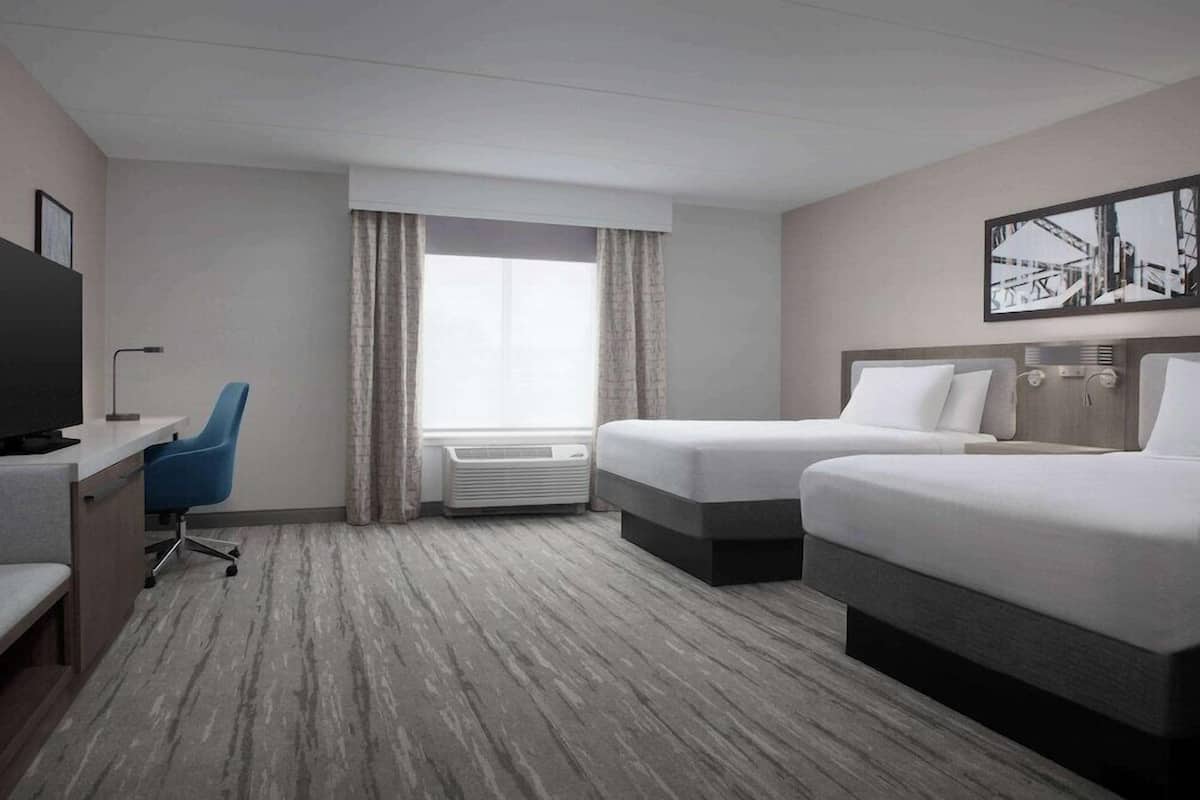 Ein Hotelzimmer mit zwei Betten, einem Schreibtisch mit Stuhl, einem Fernseher und einem großen Fenster mit Vorhängen.