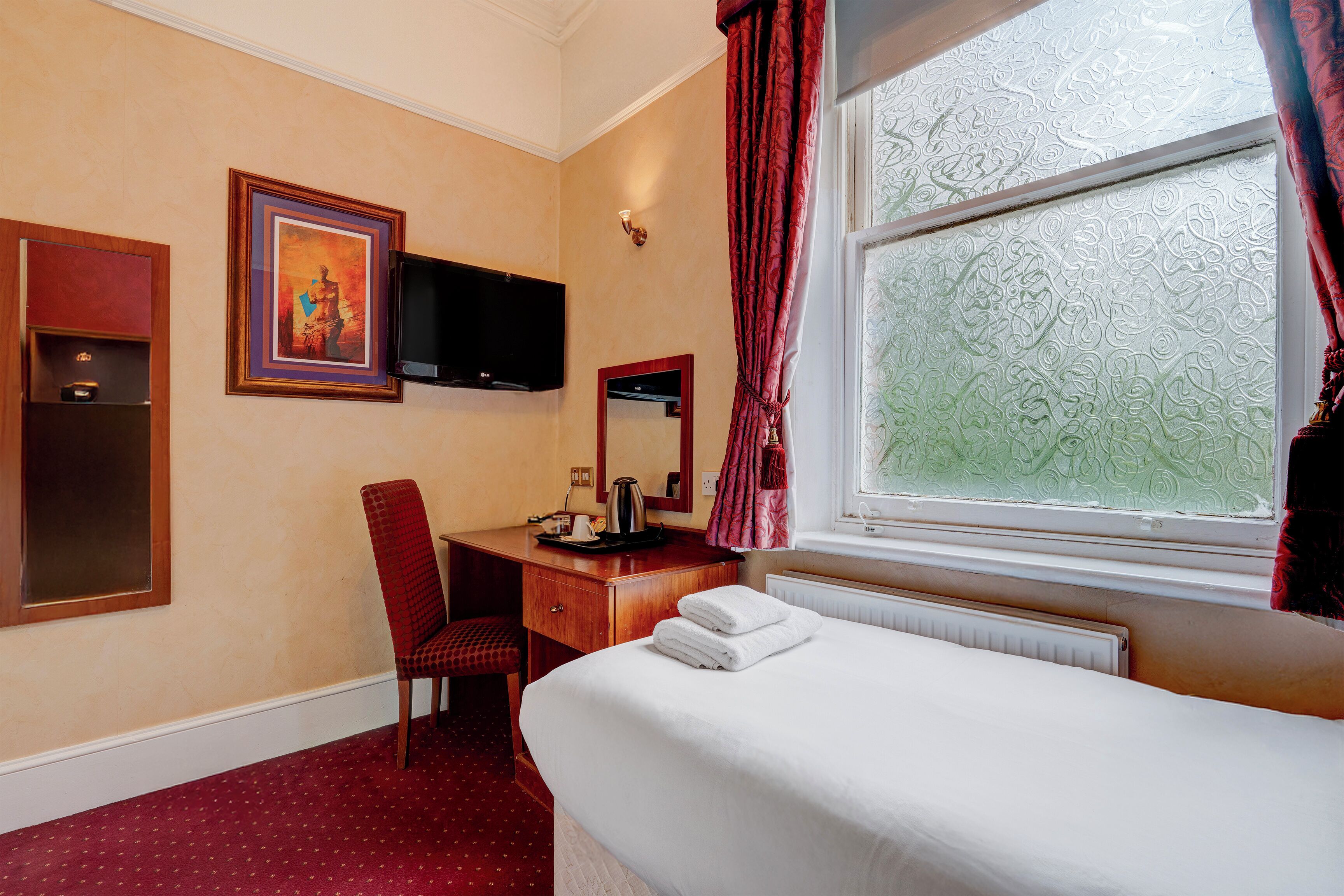 Habitación de hotel con cama, escritorio con silla, televisor, ventana con cortinas y un cuadro en la pared.