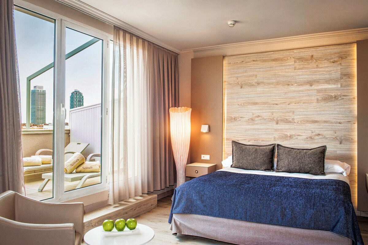 A hotel room with a large bed, a view of the city through a sliding door, a round table with apples, and a nightstand with a lamp.