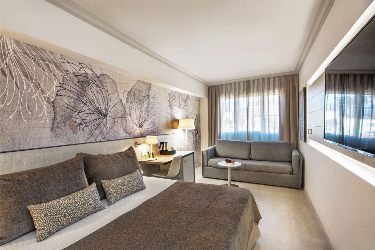 A hotel room with a large bed, a sofa, a desk, and a large abstract painting on the wall.