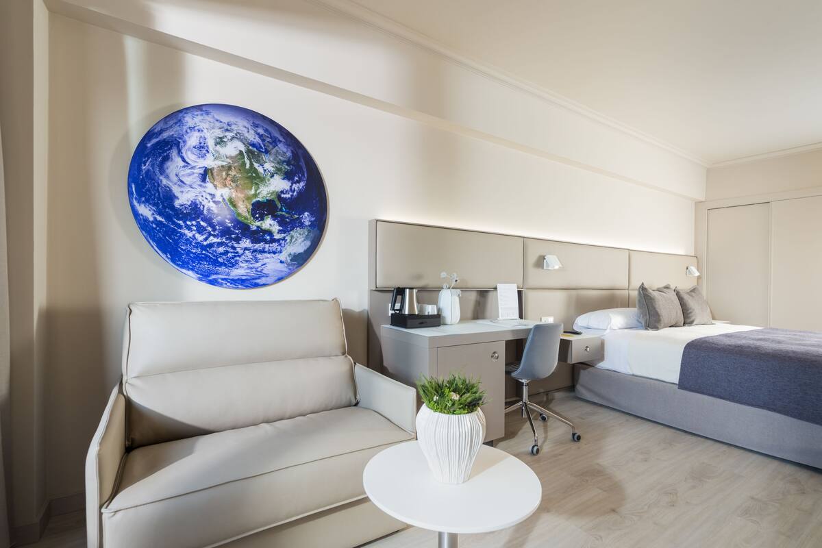 A modern hotel room with a large Earth mural, a sofa, a desk with a chair, and a bed.