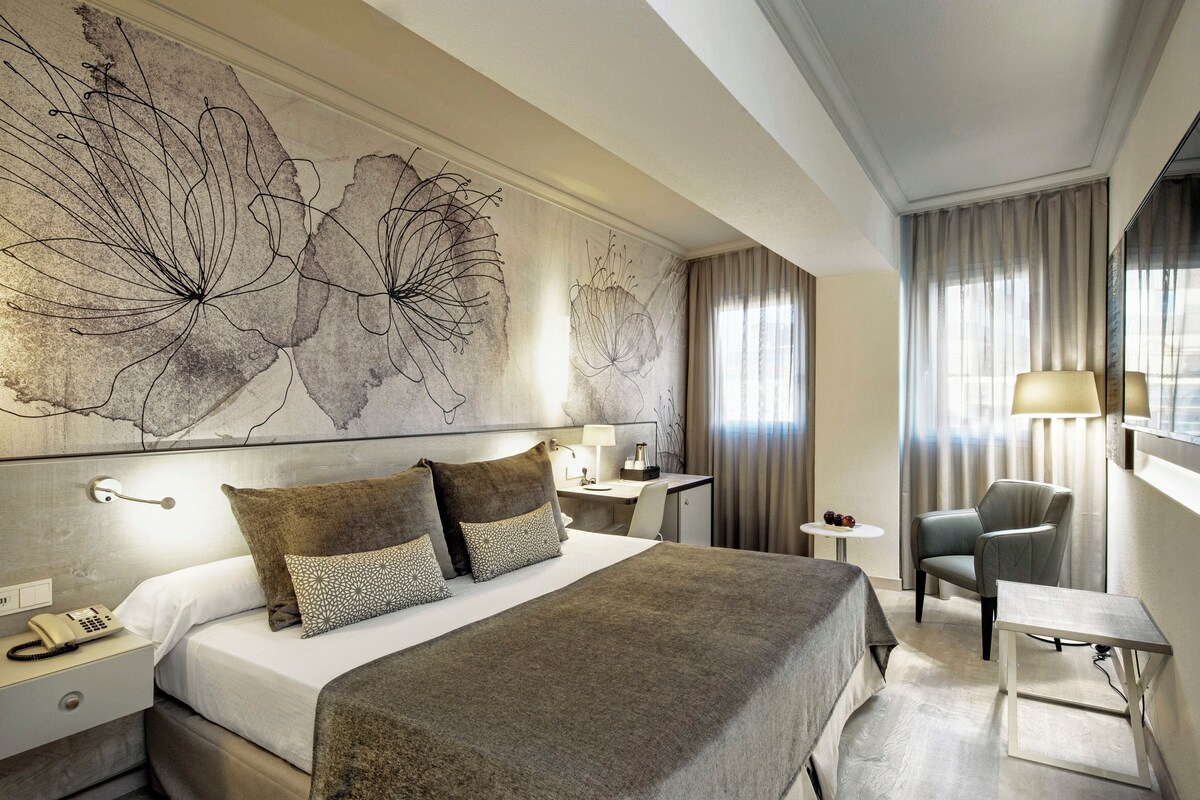 A hotel room with a large bed, a desk, a chair, a lamp, and a wall with abstract art.