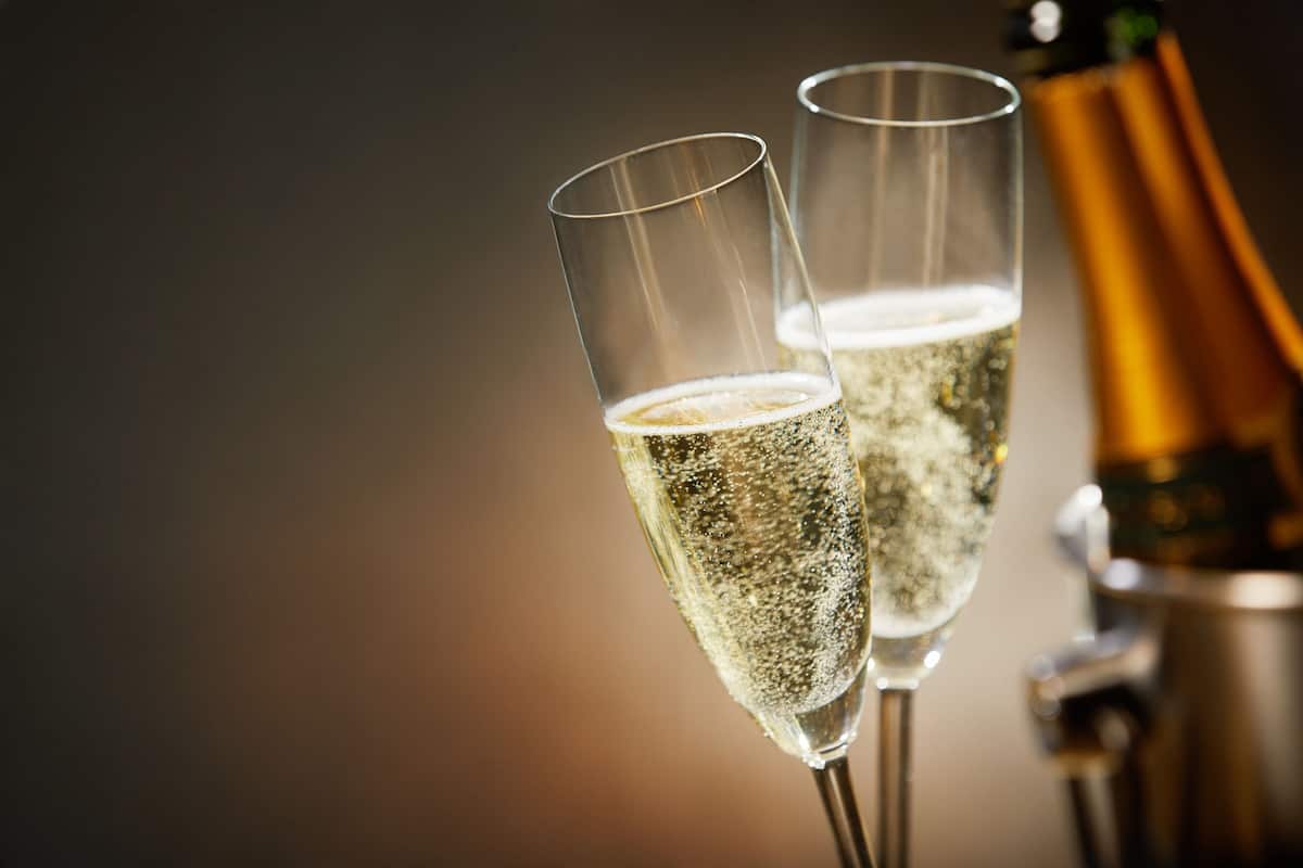 Two champagne flutes filled with bubbly, a bottle of champagne, and a gold-colored bottle in the background.