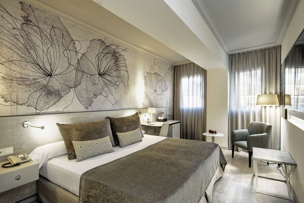 A hotel room with a large bed, a desk, a chair, a lamp, and a wall with abstract art.