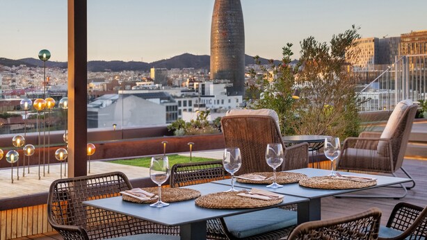 Un espacio exterior para comer con mobiliario de mimbre y una mesa dispuesta para dos, con vistas a un paisaje urbano que destaca una torre singular.
